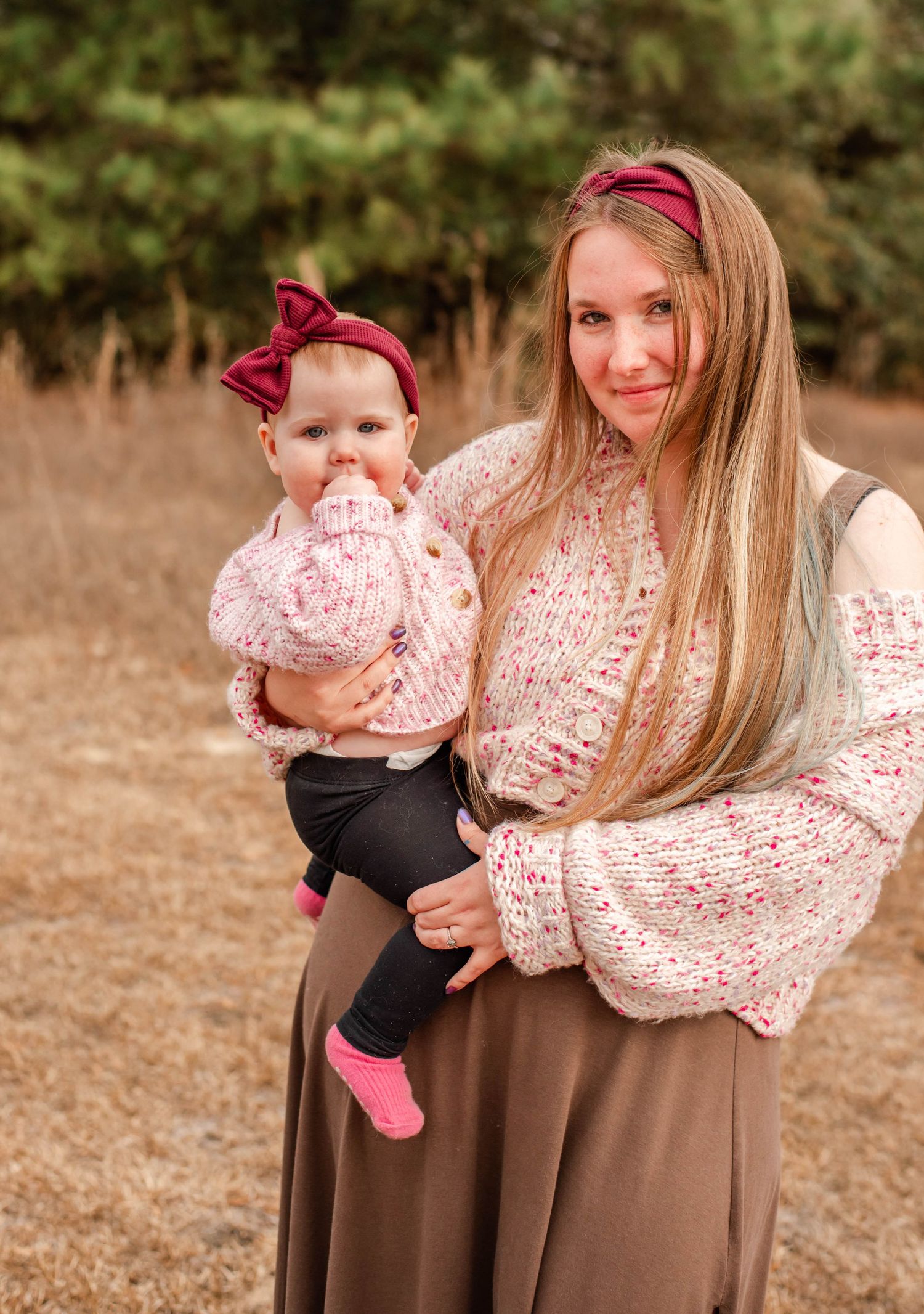 Young blonde woman holding a baby in a pink bow headband, both wearing cozy sweaters outdoors in a natural setting.