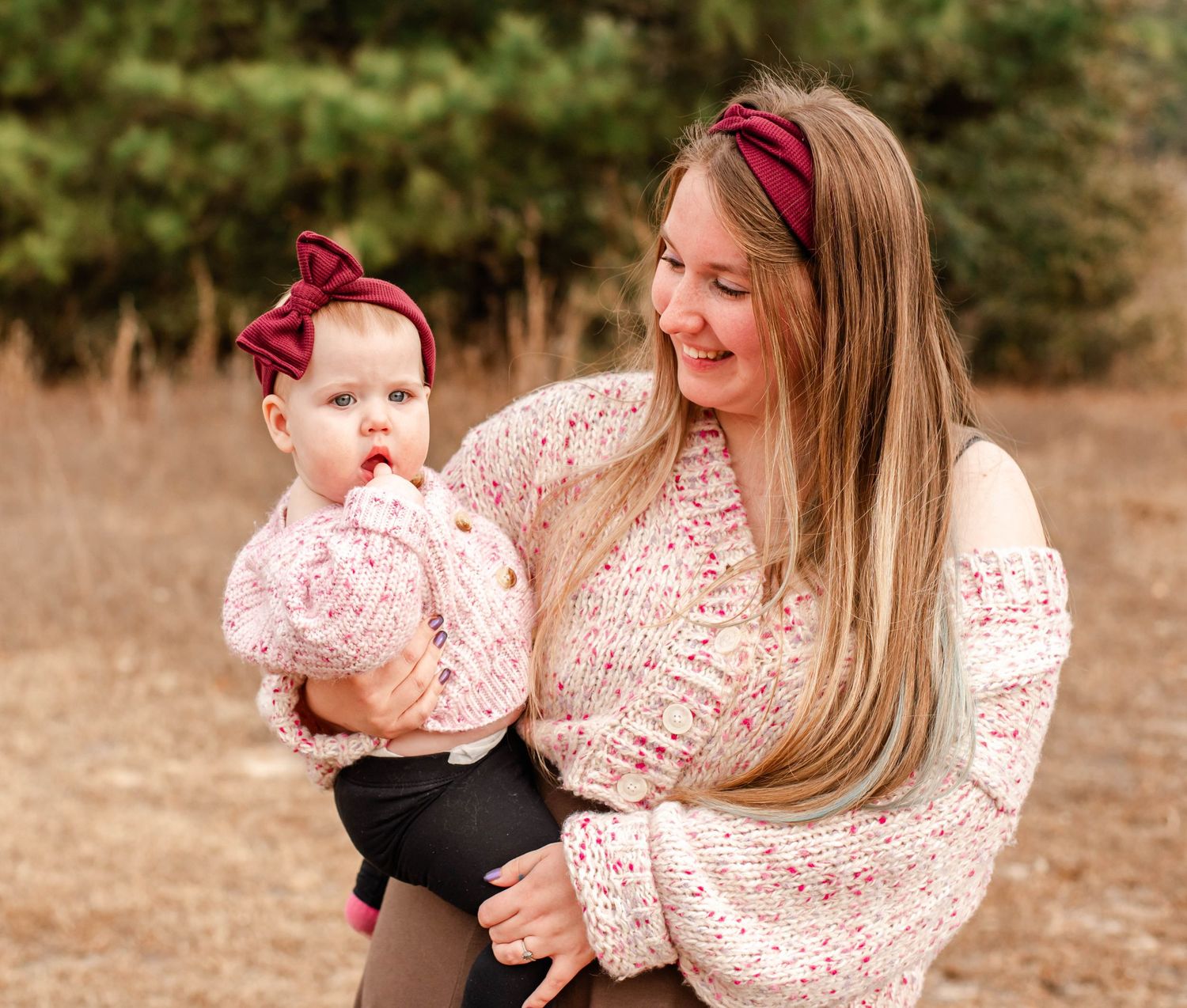 A smiling woman with long blonde hair holds a baby girl, both wearing matching pink floral outfits and burgundy bow headbands outdoors.
