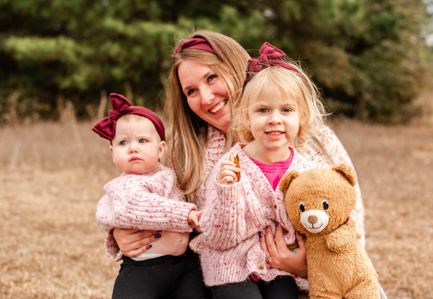 A smiling mother holds two young daughters in matching pink outfits and burgundy bows outdoors, one child holding a teddy bear.