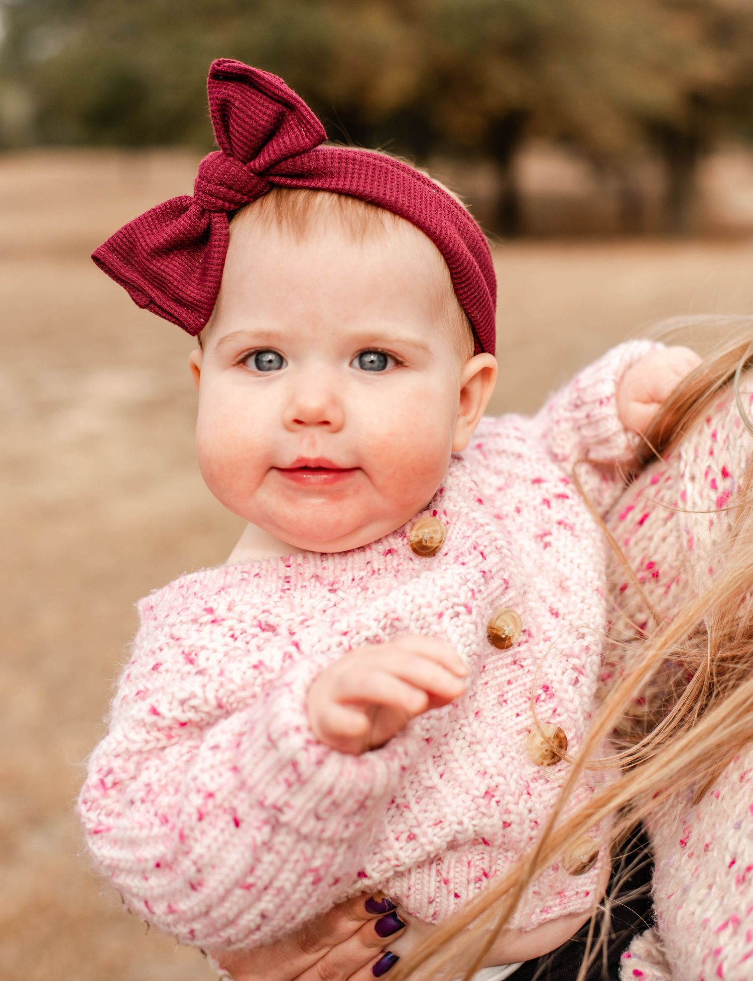 Adorable baby girl wearing a pink knit sweater and burgundy bow headband, being held outdoors in a park setting.