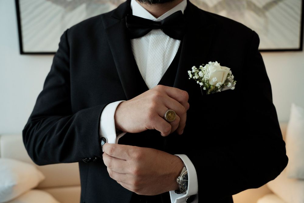 Groom in black tuxedo with bow tie and white rose boutonniere adjusting cufflinks before wedding ceremony.