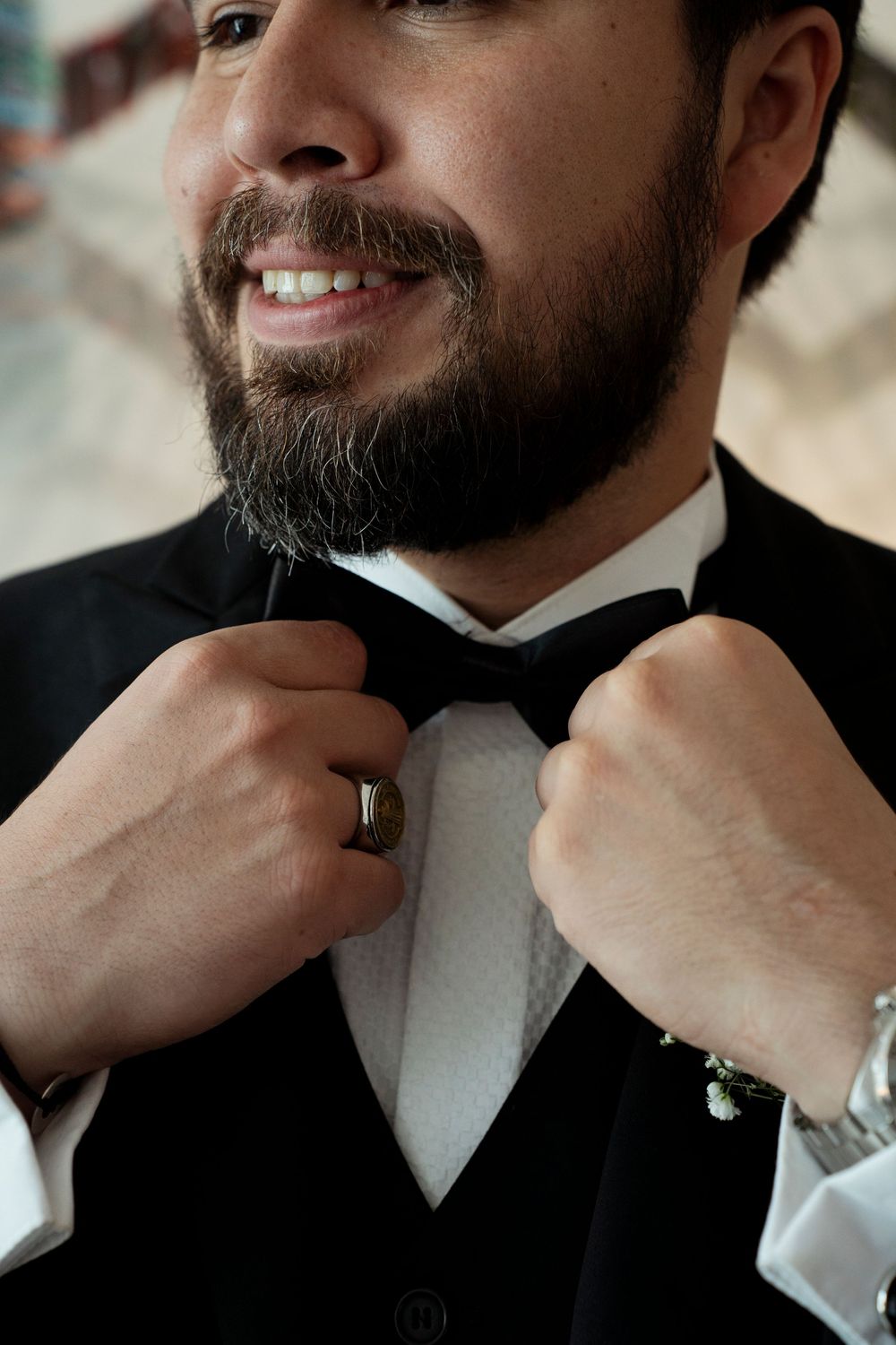 Smiling groom in black tuxedo and bow tie adjusting his neckwear, wearing a wedding ring on his finger.