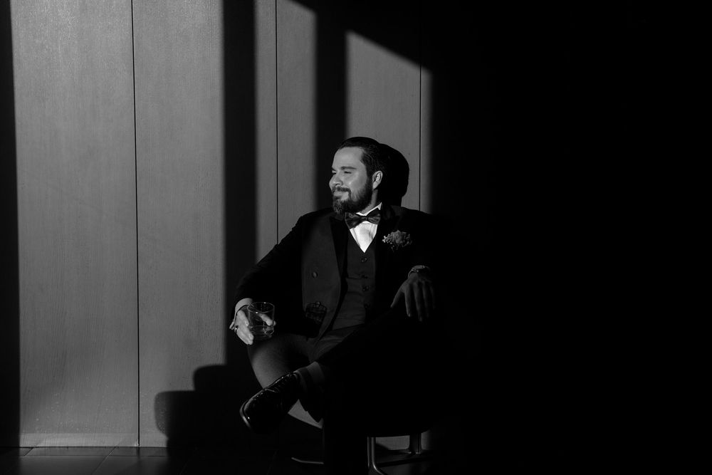Black and white portrait of a bearded groom in a suit sitting in a chair, bathed in dramatic light and shadow.