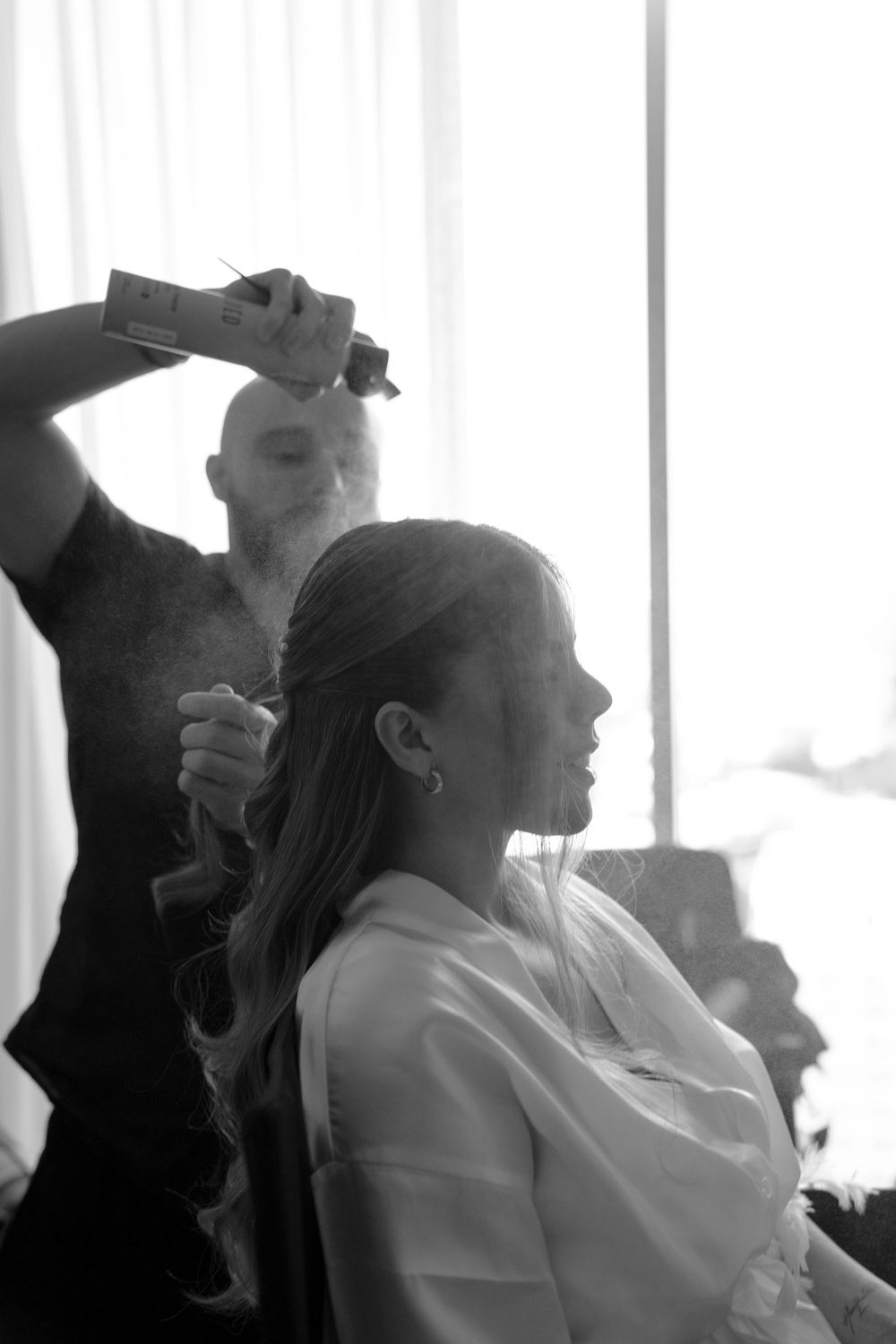 Black and white photo of a hairstylist spraying hairspray on a bride with long curly hair during wedding preparations.