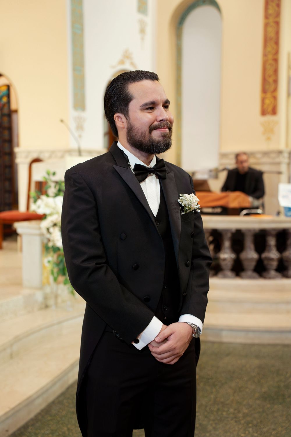 Smiling groom in black tuxedo with boutonniere stands at church altar awaiting bride during wedding ceremony.