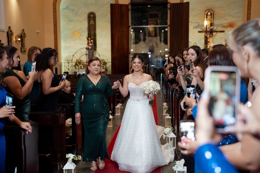 Bride walks down church aisle in white gown with mother in green dress, guests capturing photos on smartphones.