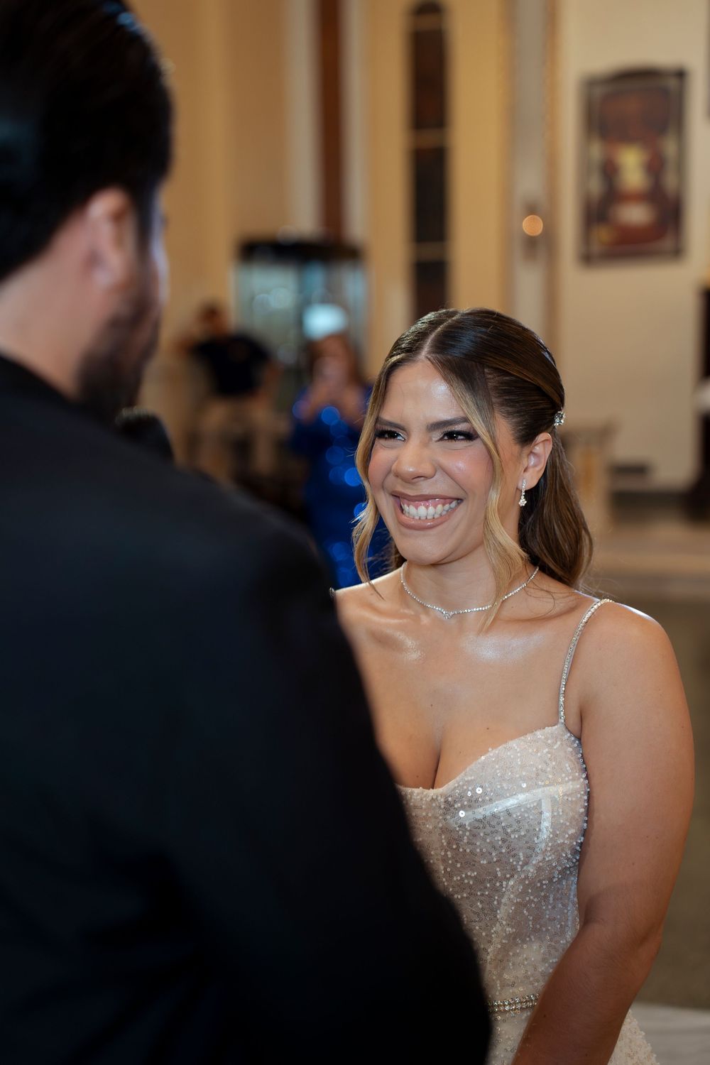 Bride in beaded gown smiling joyfully at groom during wedding ceremony indoors.