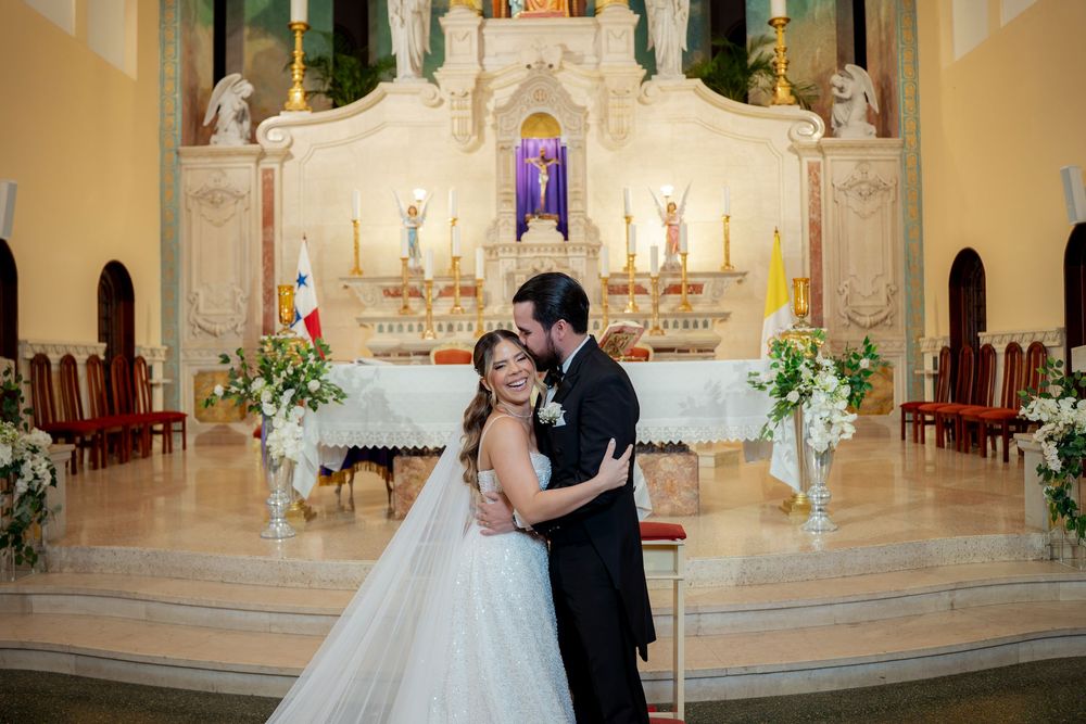 Bride and groom share romantic embrace at church altar during wedding ceremony with white floral decorations.