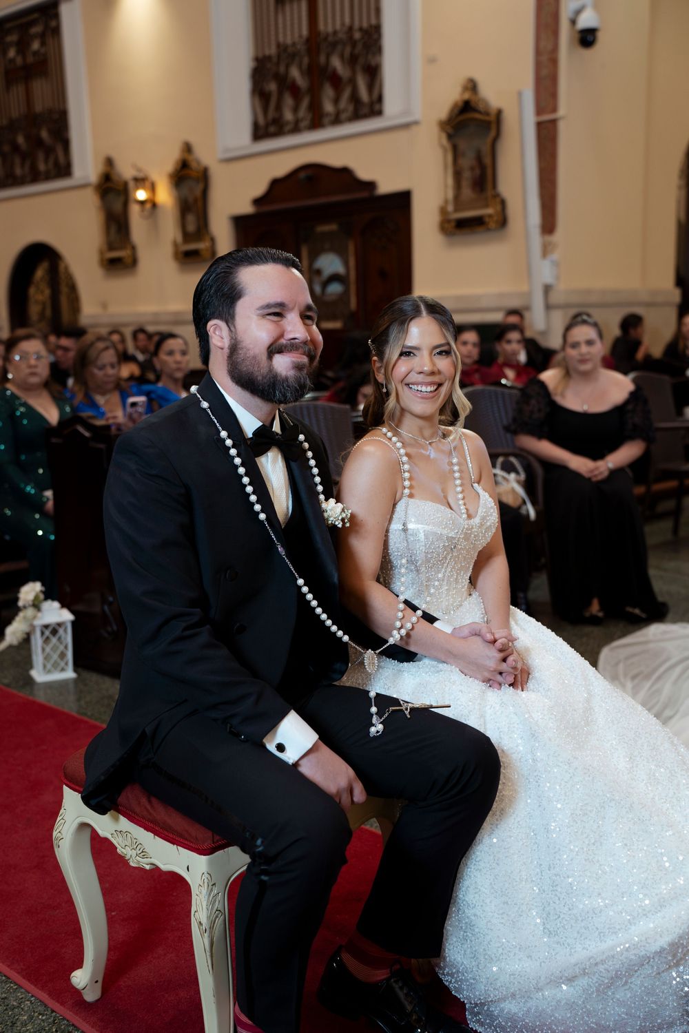 Smiling bride and groom sitting together during a church wedding ceremony, wearing formal attire with pearl accessories.