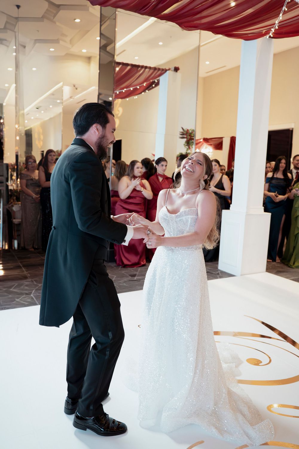 Bride and groom share their first dance, laughing joyfully on a white dance floor surrounded by wedding guests.