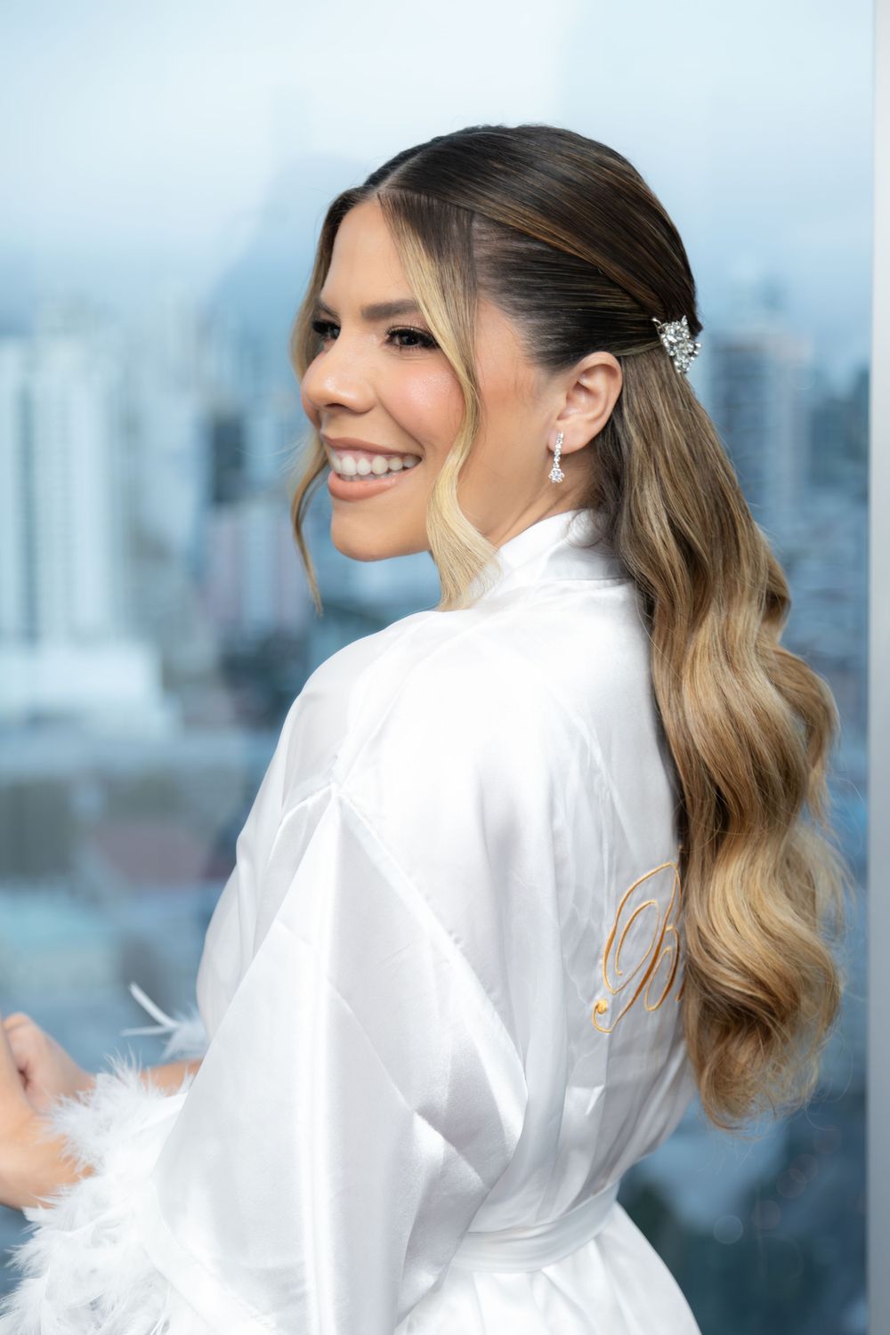 Smiling bride in white satin robe with feather trim, wearing pearl earrings and crystal hair clip, long wavy ombre hair.
