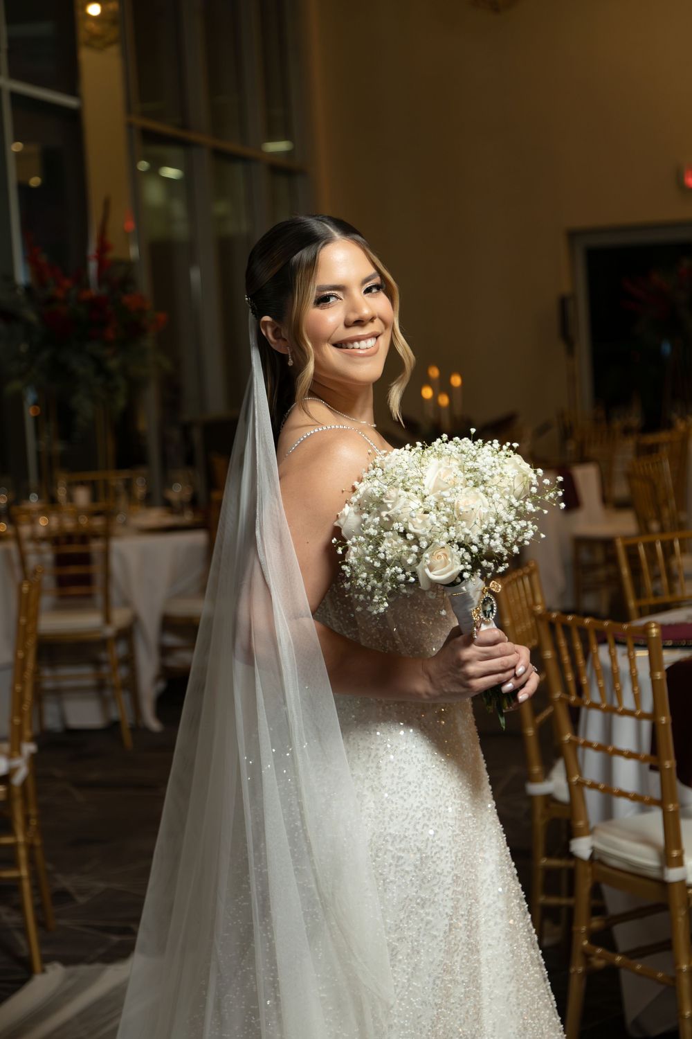 Smiling bride in a sparkly white gown and long veil holds a baby's breath bouquet in an elegant gold-chaired reception hall.