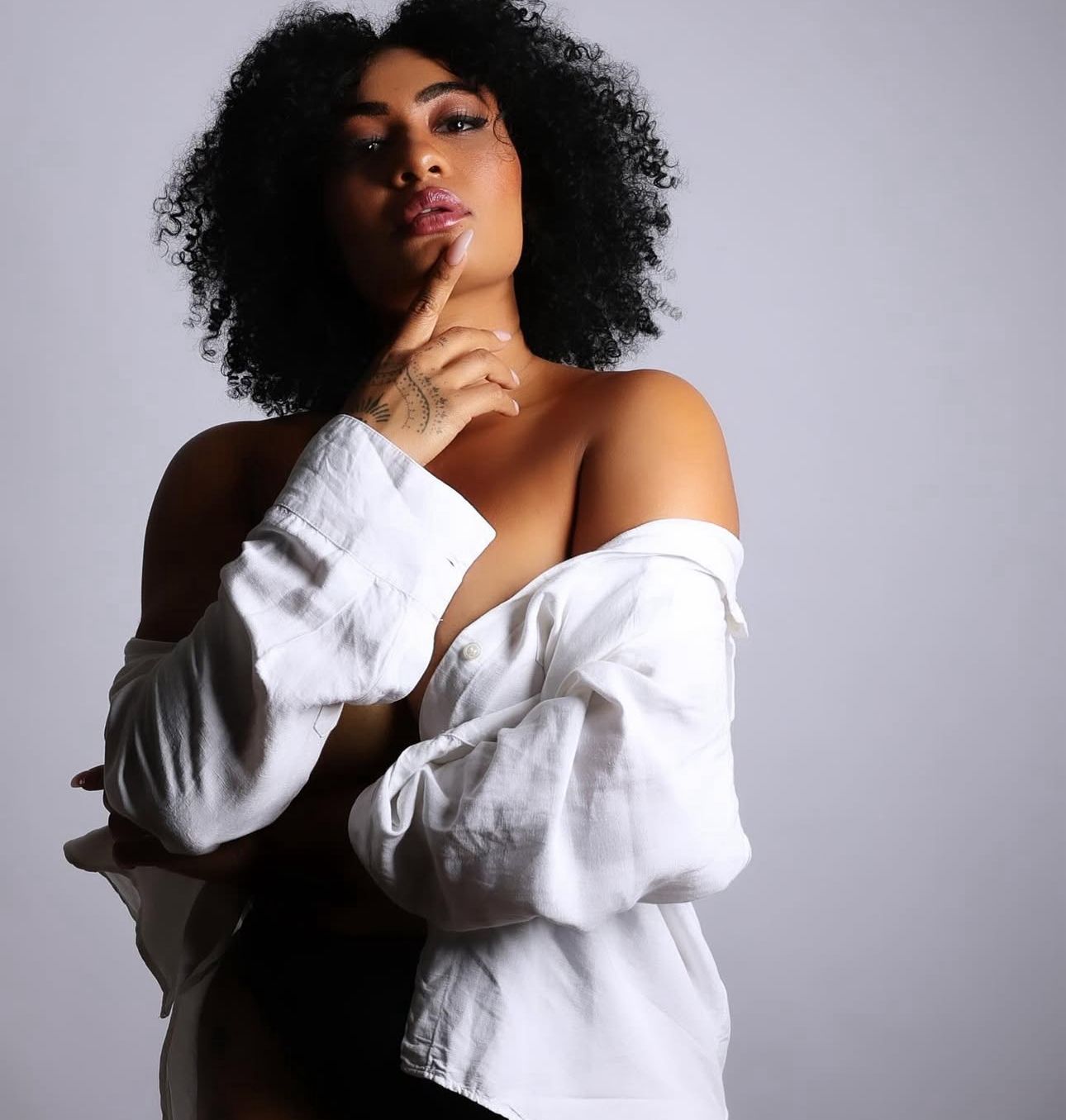 A woman with curly hair poses confidently in an off-shoulder white shirt against a light gray background.