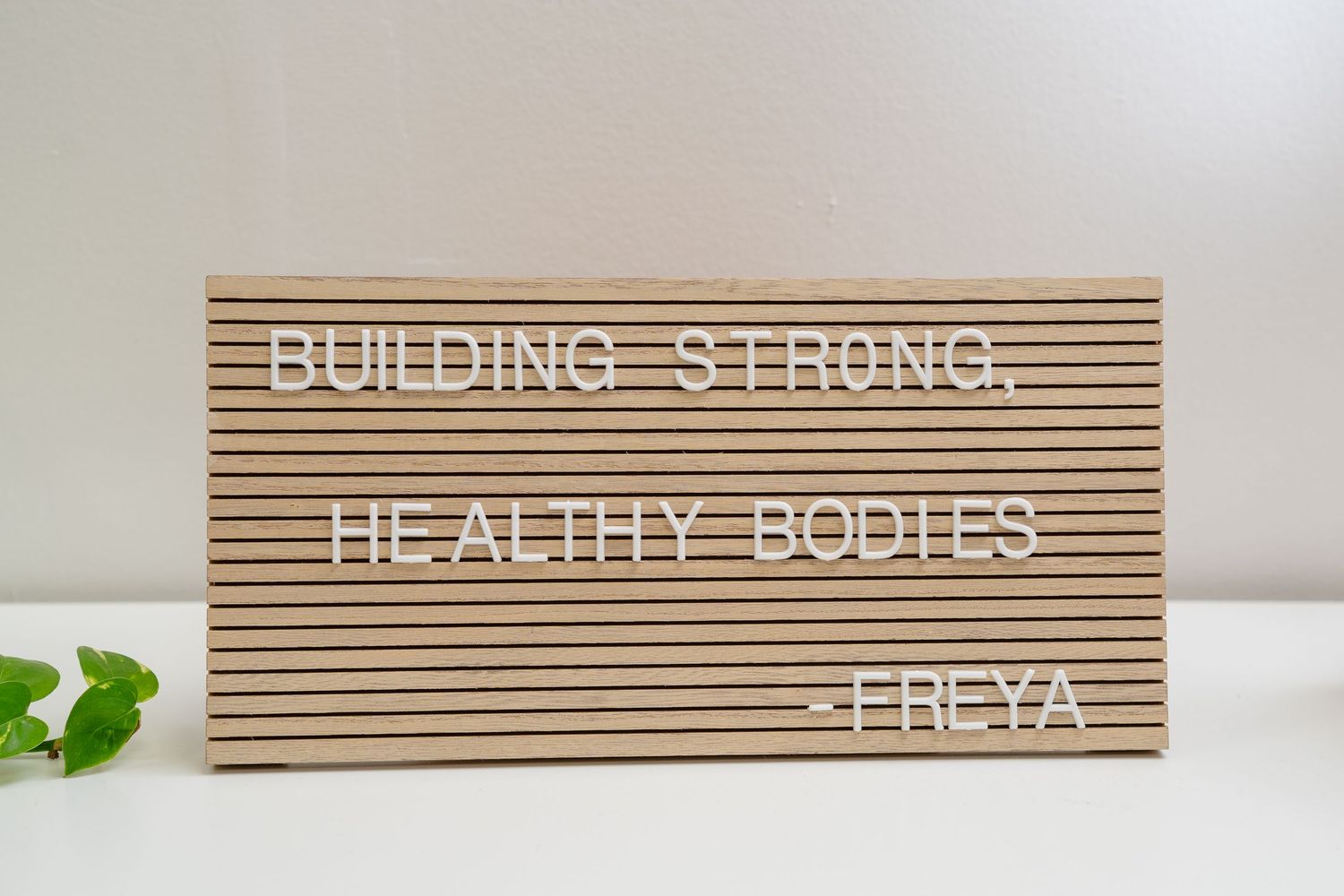 Wooden letter board displaying 'Building Strong, Healthy Bodies -Freya' motivational fitness quote on white background.