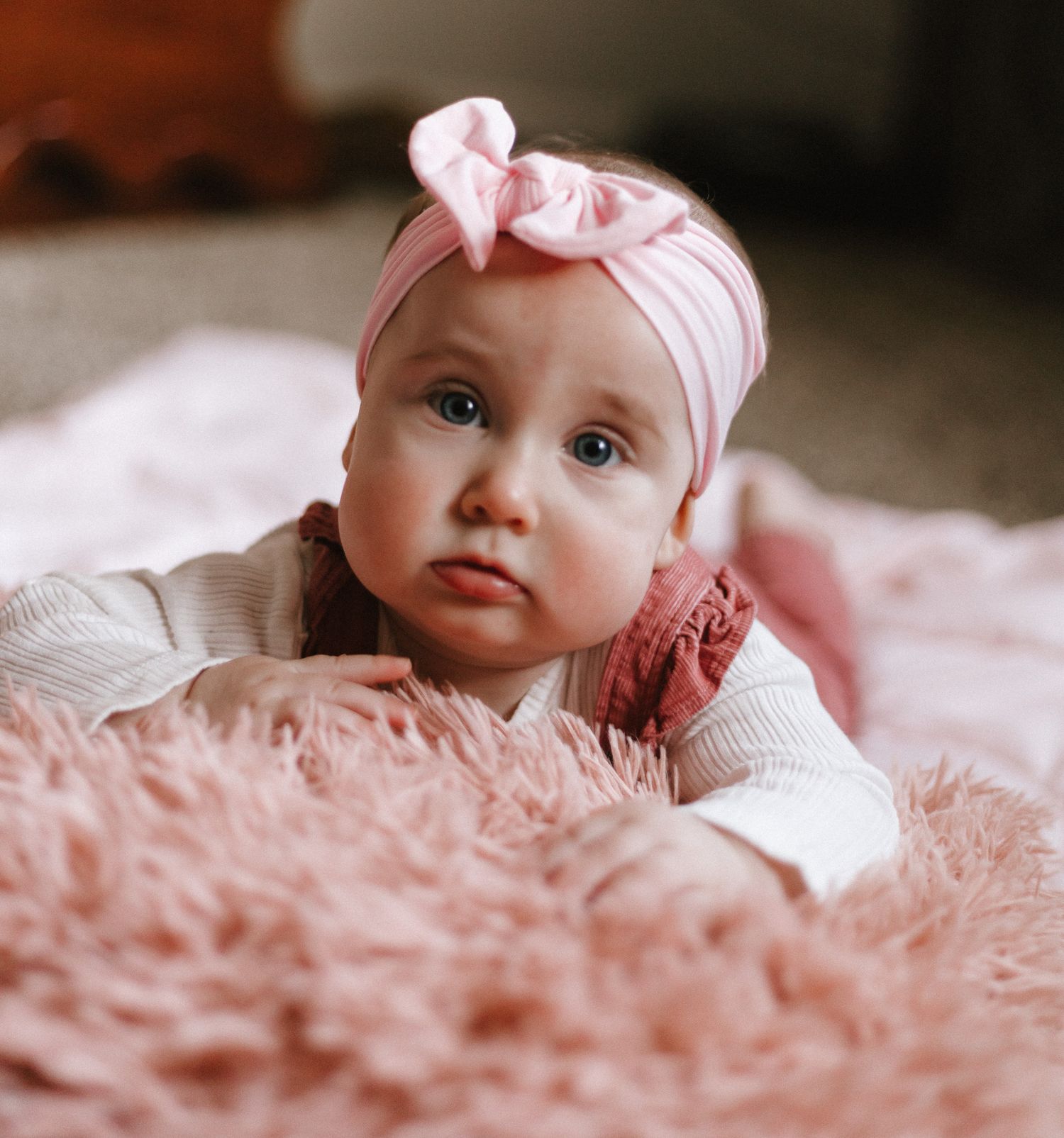 Adorable baby girl with pink bow headband lying on fluffy pink rug.
