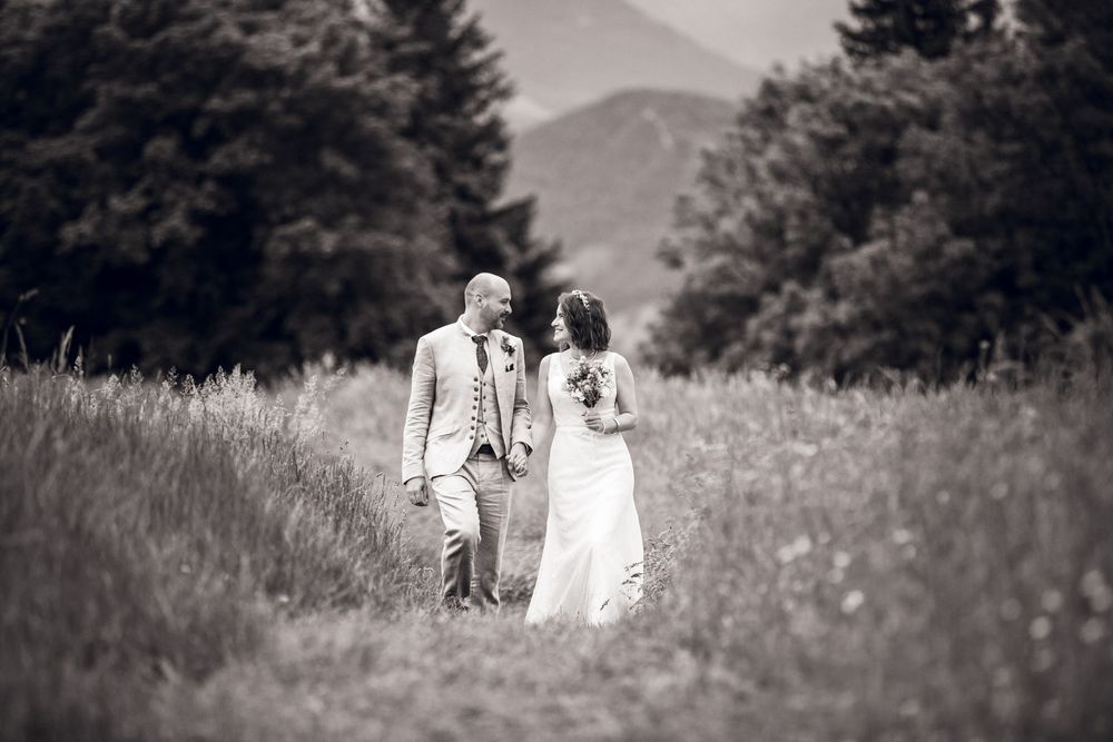 Black and white photo of a bride and groom walking through a meadow with mountains and trees in the background.
