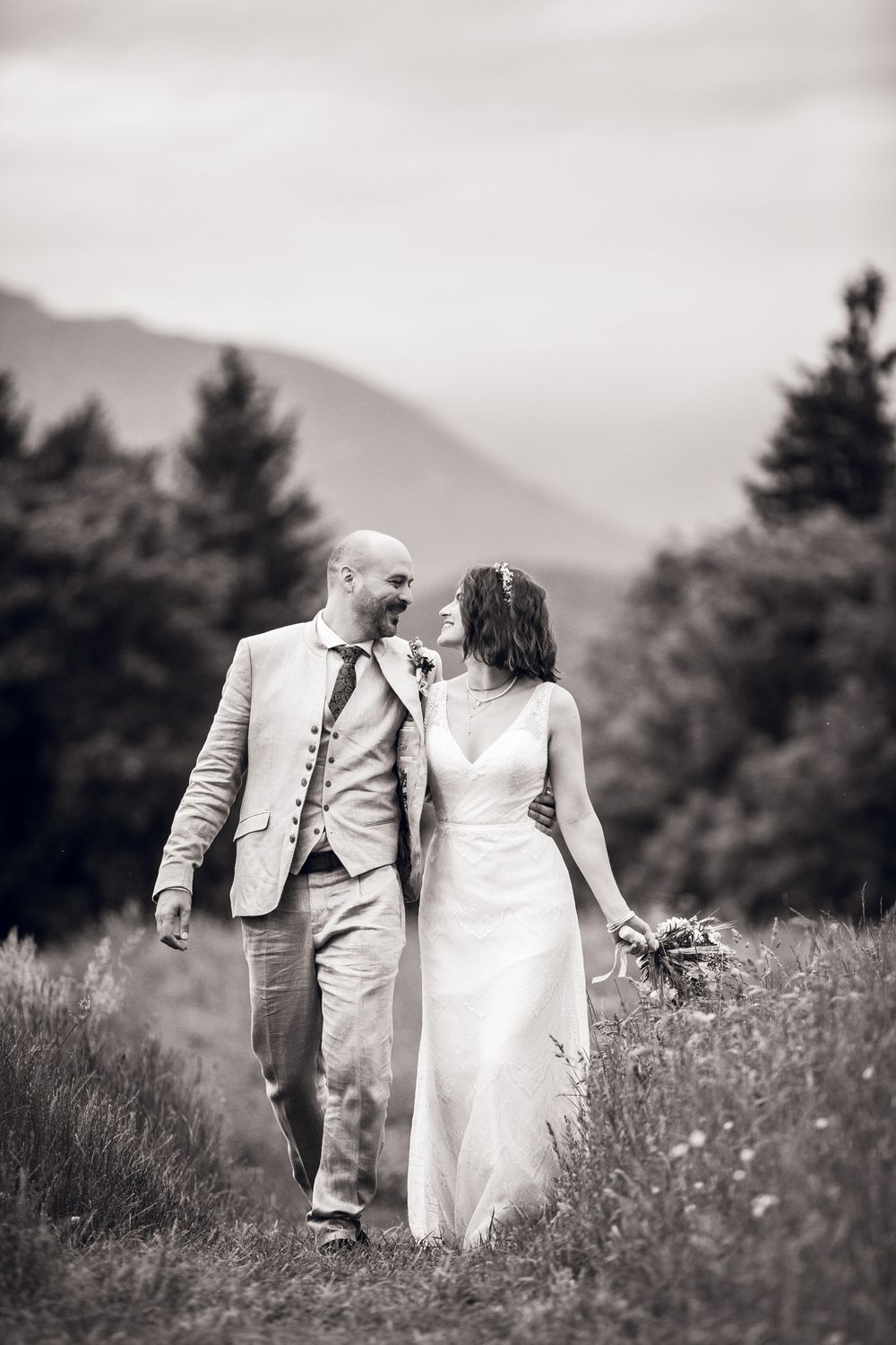 Black and white photo of a bride and groom walking through a mountain meadow, smiling at each other on their wedding day.