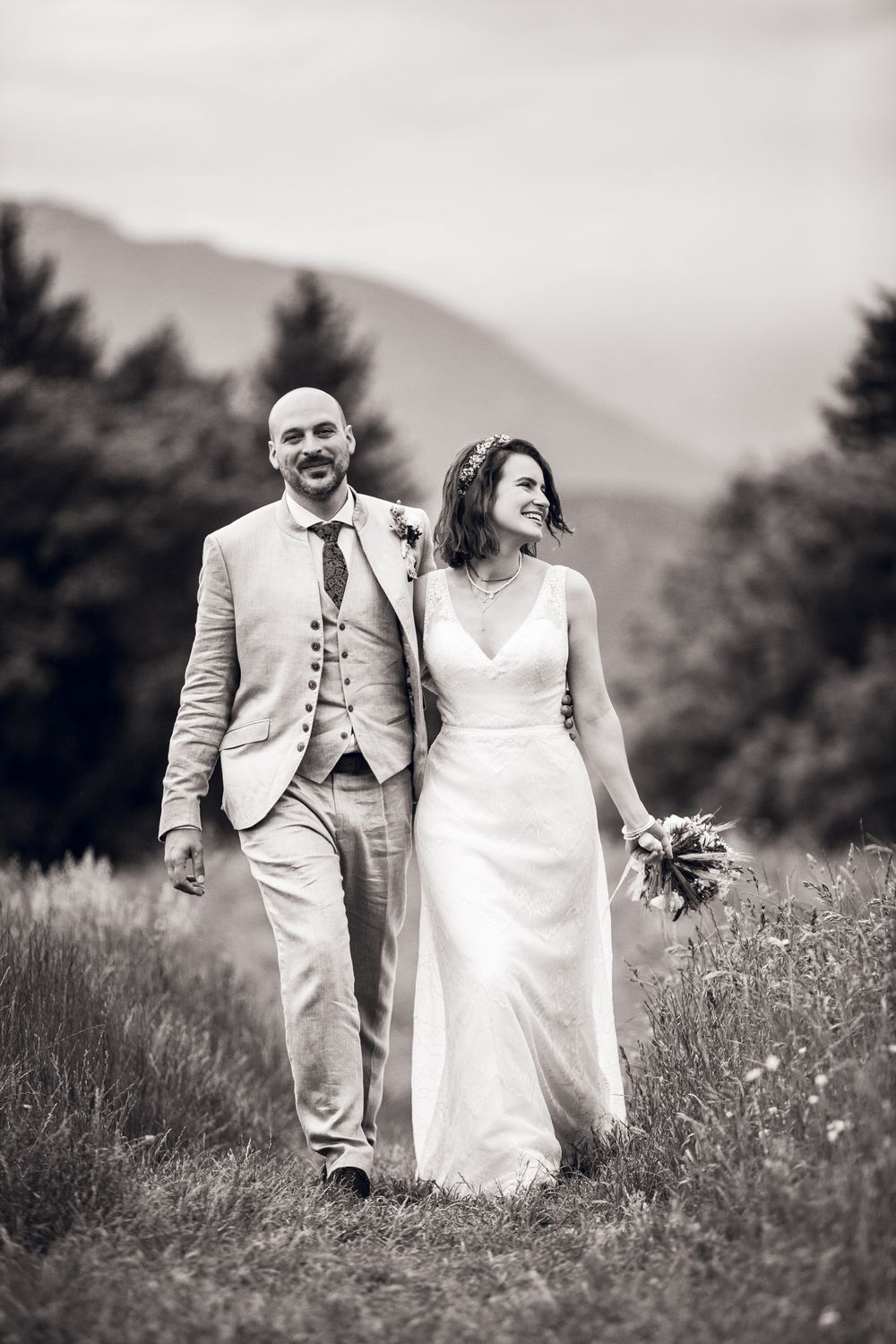 Bride and groom walk through meadow with mountain backdrop in black and white wedding portrait.