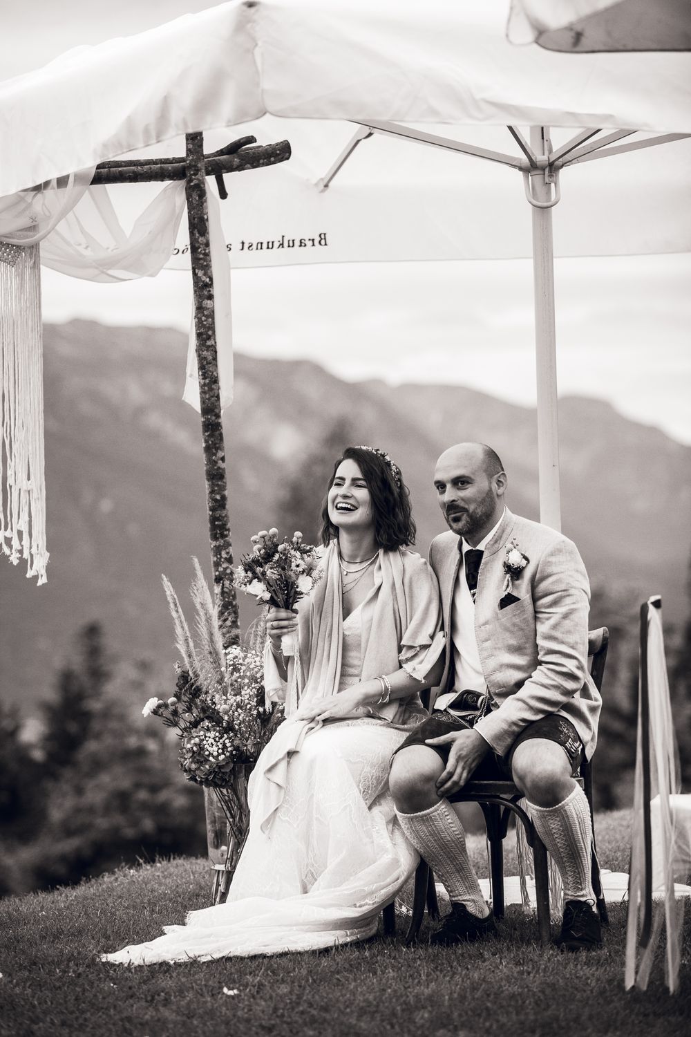 Black and white photo of bride and groom laughing together seated under a canopy with mountain views.