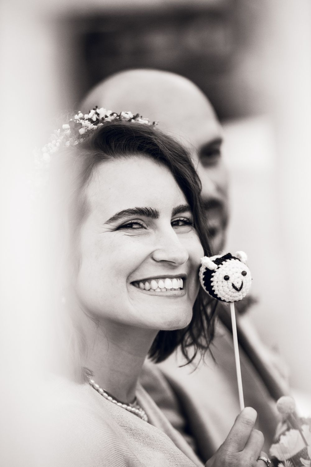Smiling bride with floral crown holds a decorative cake pop, with bald groom behind her, in black and white wedding photo.