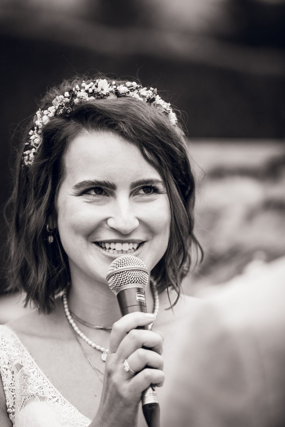 Smiling bride wearing floral crown speaks into microphone during outdoor wedding ceremony in black and white.