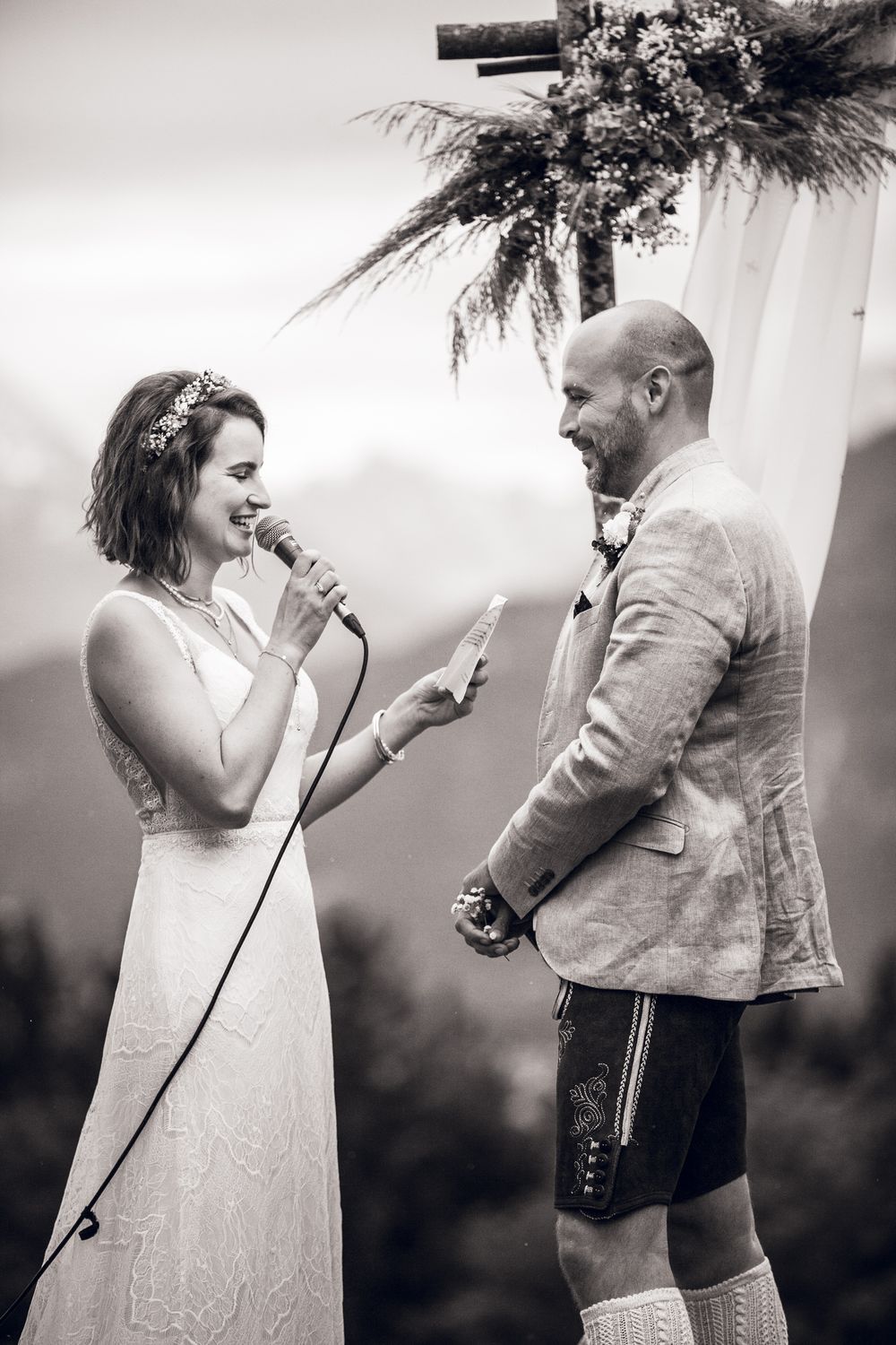Bride reads vows into microphone while groom smiles at outdoor mountain wedding ceremony.
