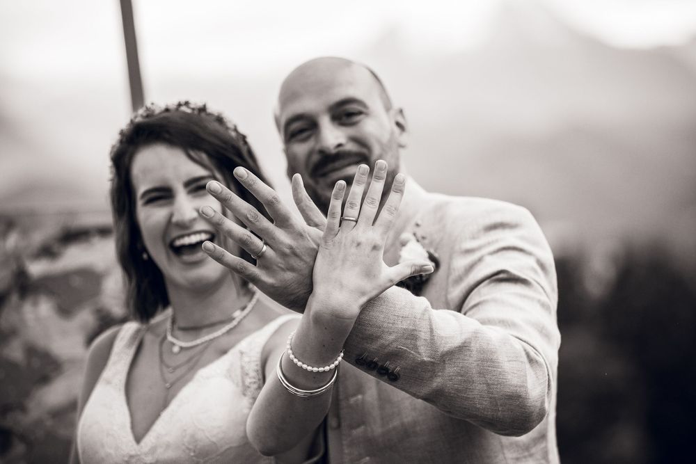 Joyful bride and groom showing off their wedding rings to camera in black and white photo.