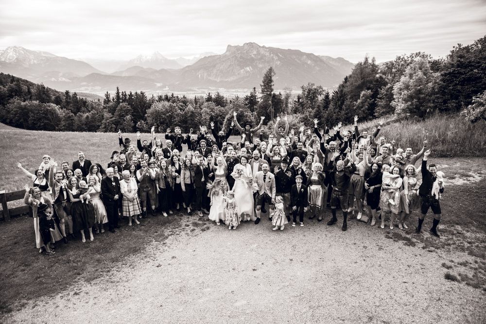 Black and white aerial group photo of wedding guests celebrating outdoors with mountain backdrop.
