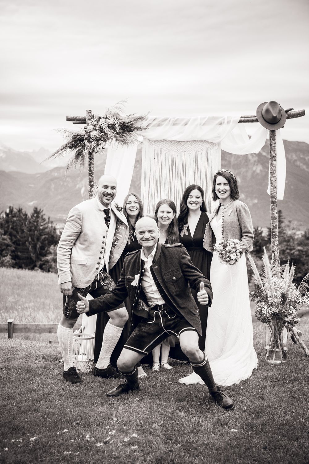 Black and white wedding photo of bride, groom, and guests posing joyfully before a floral arch with mountain backdrop.