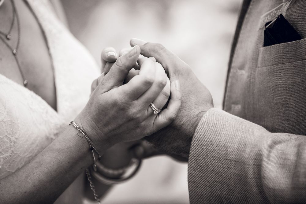 Black and white close-up of bride and groom holding hands showing wedding rings and bracelets.
