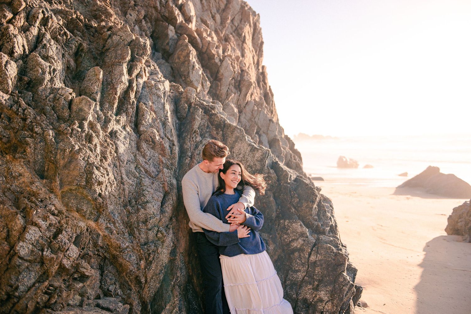 Couple embracing tenderly against rocky coastal cliffs on a sandy beach, sharing an intimate moment at golden hour.