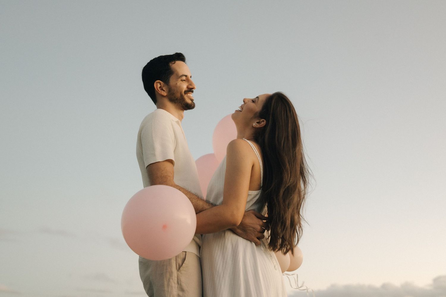 Couple sharing romantic kiss outdoors surrounded by pastel balloons at scenic overlook.