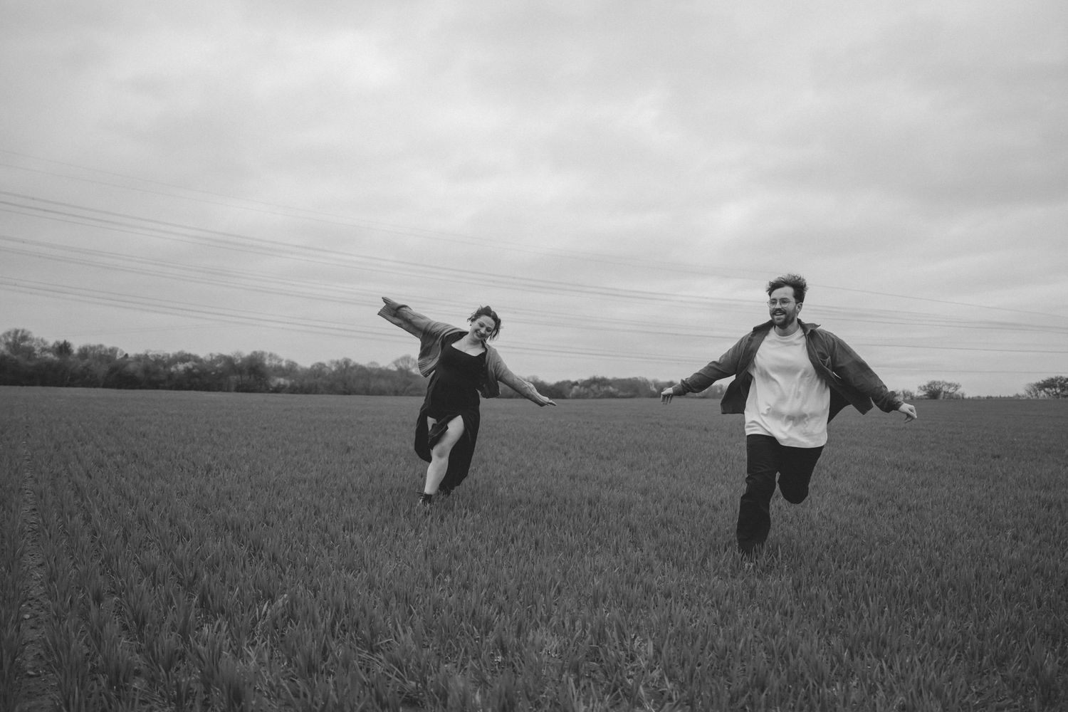 Two newly engaged people joyfully running through a vast green field on a cloudy day near Bristol in a black and white photo.