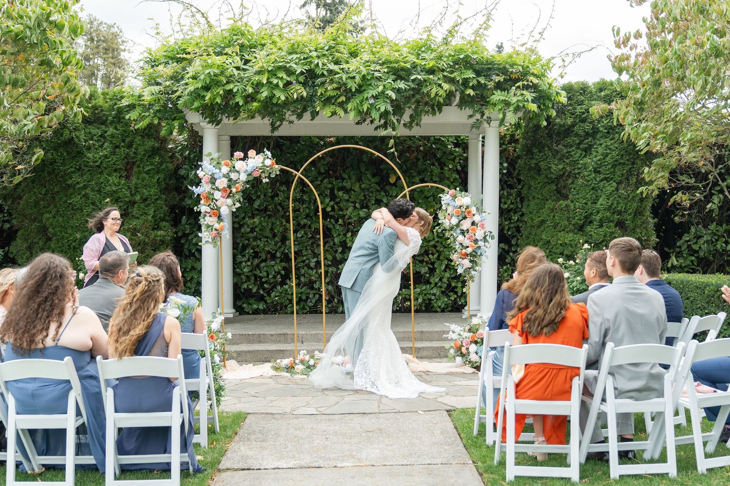 Bride and groom share first kiss at outdoor wedding ceremony beneath golden floral arch with white pergola backdrop.