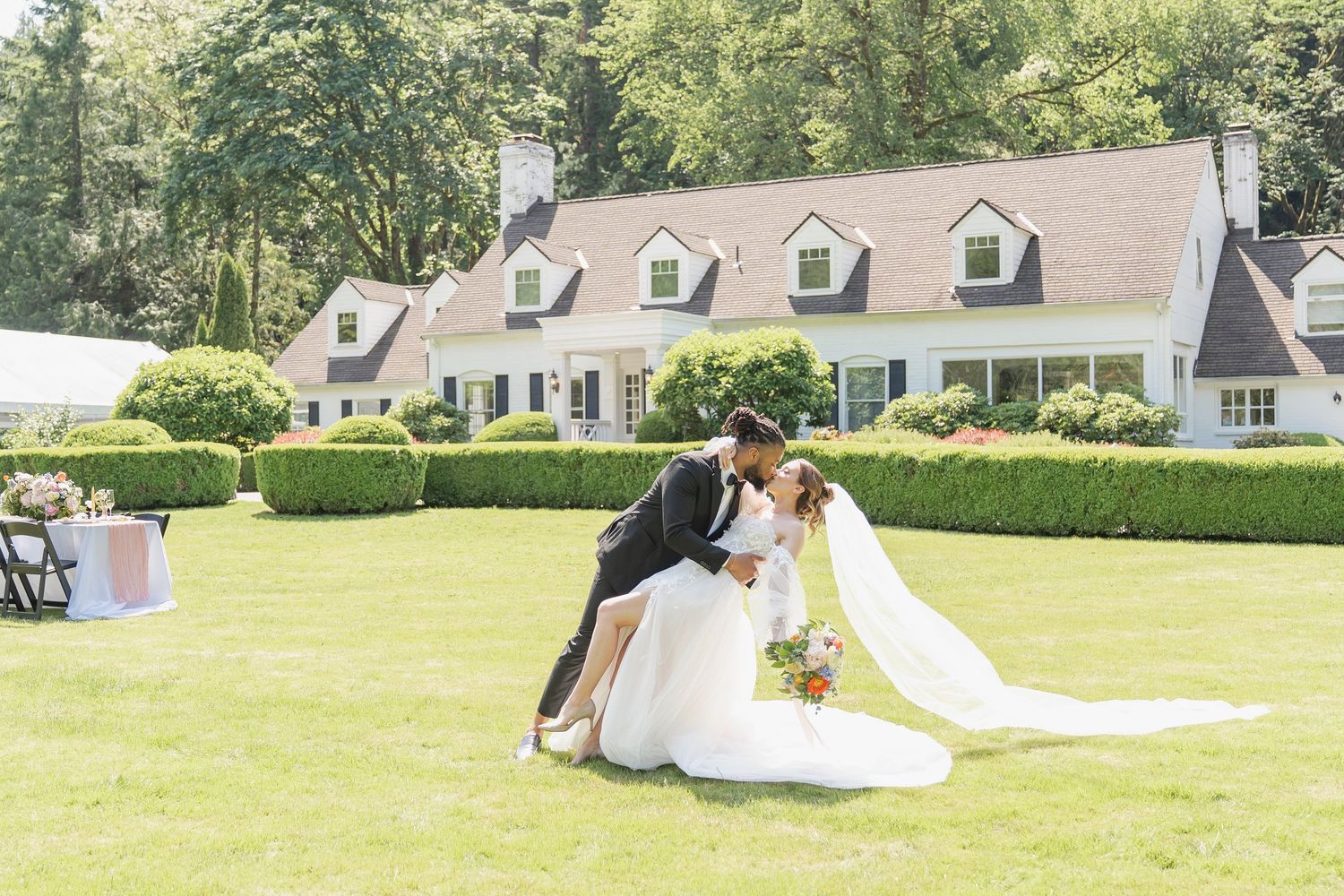 Bride and groom share a romantic dip kiss on lush green lawn outside elegant white country estate wedding venue.