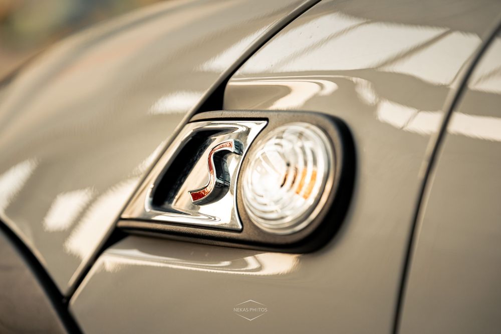 Close-up of a MINI Cooper S badge and headlight on a sleek silver car hood in natural sunlight.