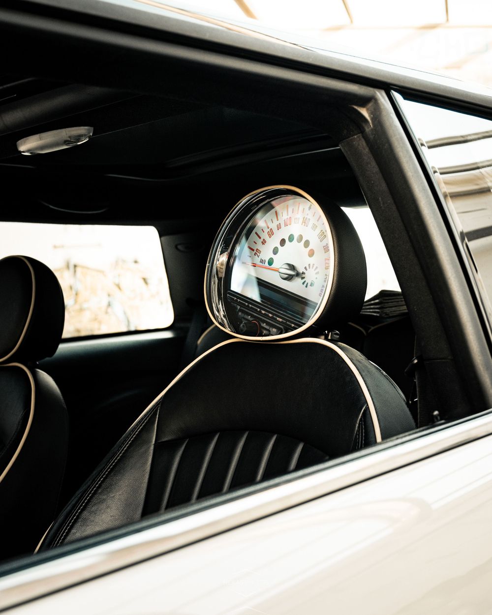 Interior view of a MINI Cooper showing black leather seats with gold piping and a circular speedometer mounted on the headrest.