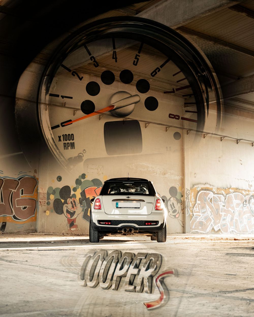 A MINI Cooper S viewed from behind in an urban setting with graffiti walls and a large tachometer backdrop.