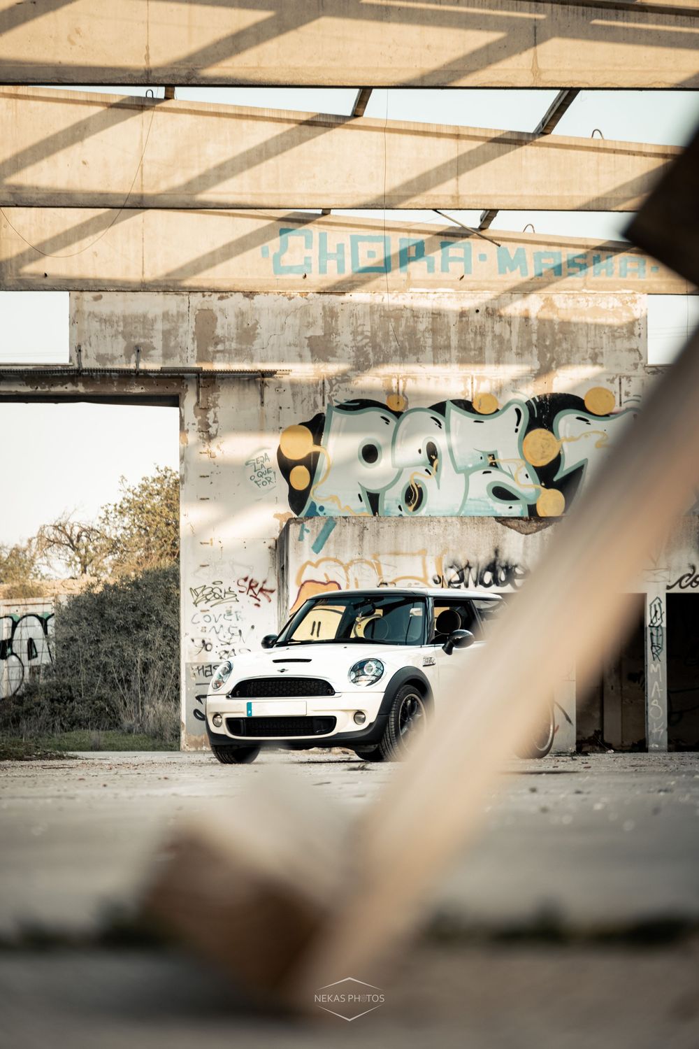 White Mini Cooper parked in an abandoned building with colorful graffiti walls, viewed through a wooden frame.