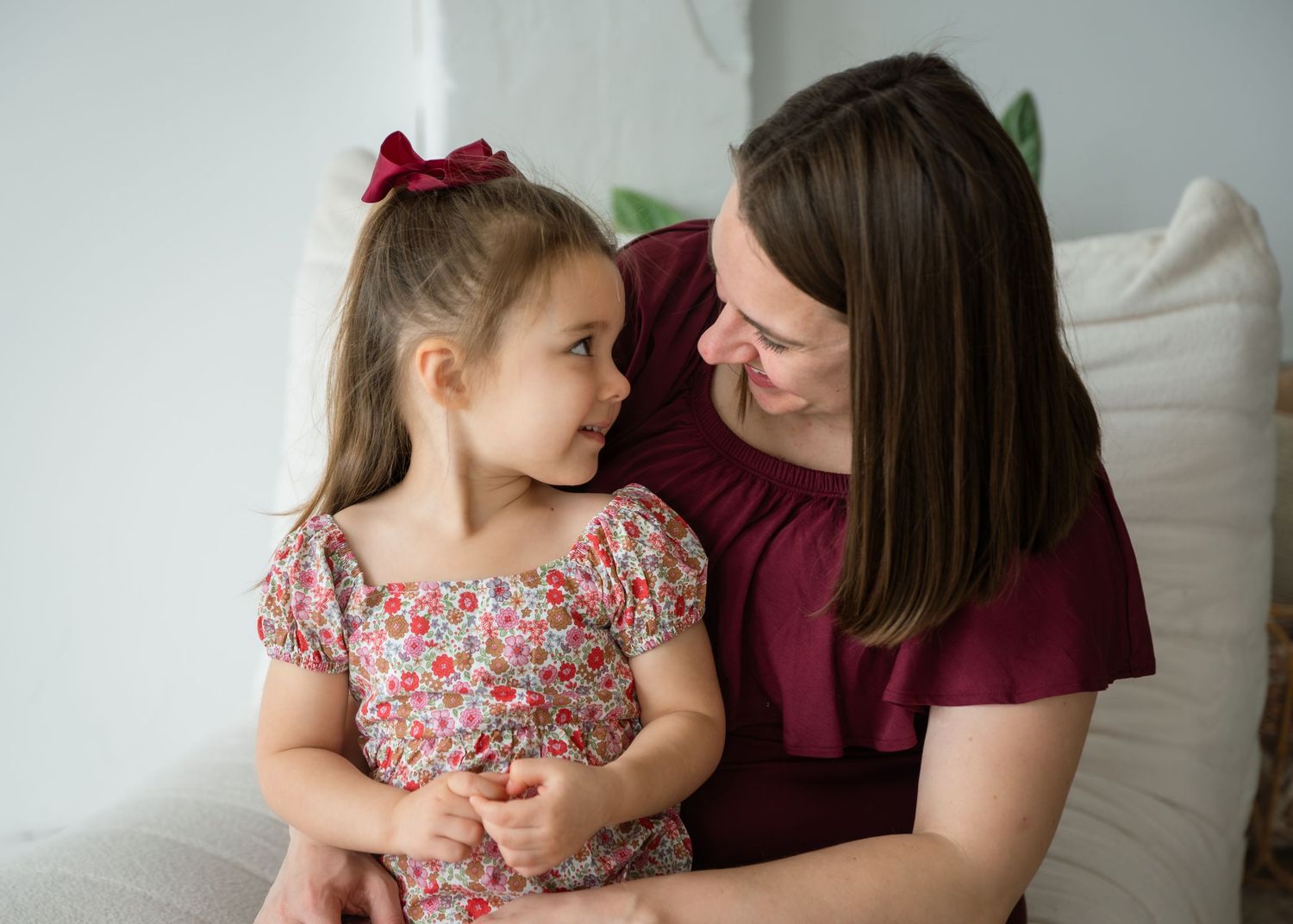 A toddler with a pacifier sits beside a pregnant woman in a burgundy dress on a beige sofa.