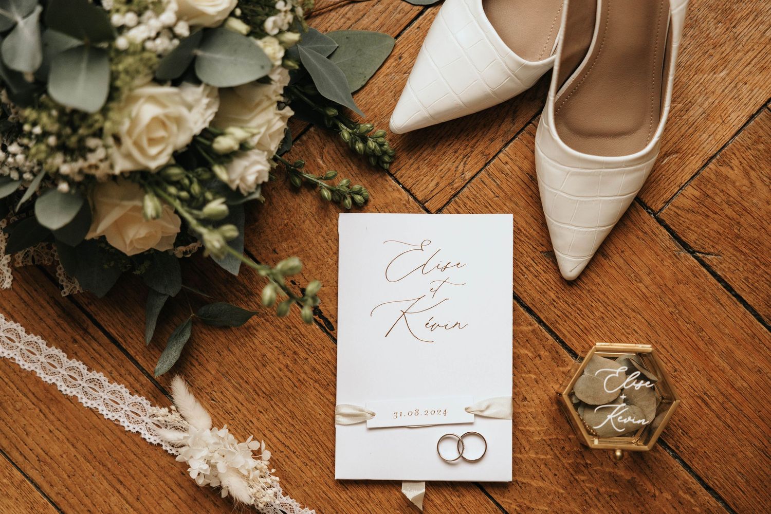 Elegant wedding flat lay featuring white heels, bridal bouquet, invitation, rings, and pearl jewelry on rustic wood floor.