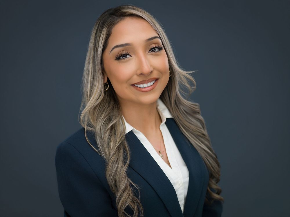Professional headshot of a young woman in a navy blazer with long highlighted hair, smiling confidently.