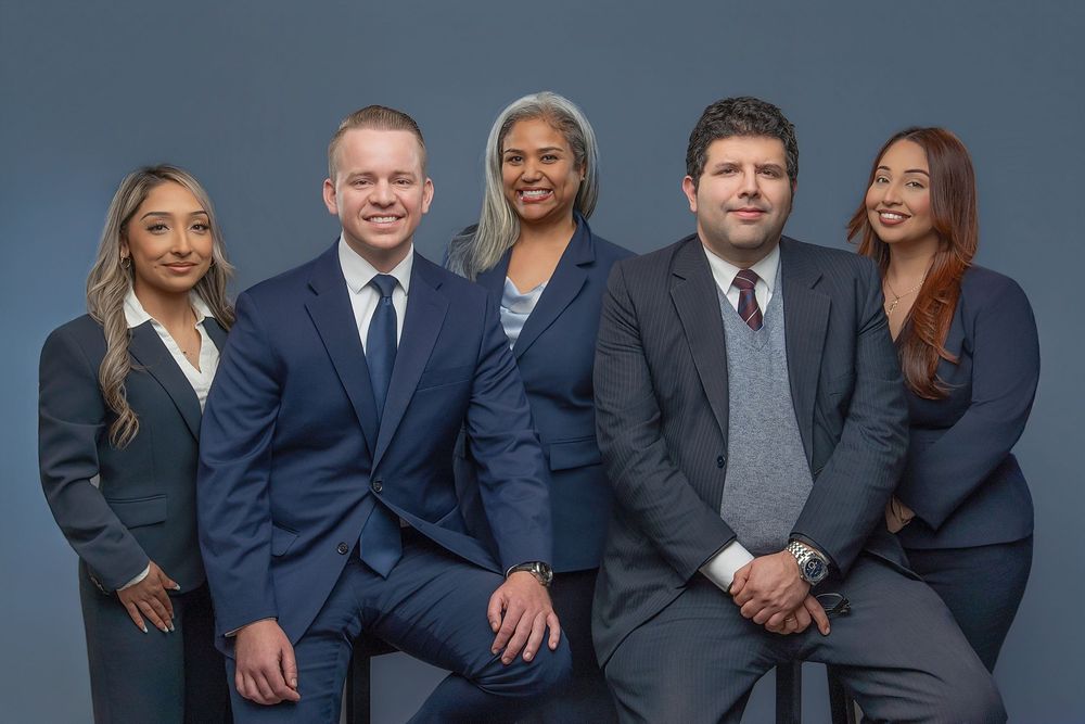 Five professional attorneys in navy suits pose together for a formal law firm group portrait against a gray background.