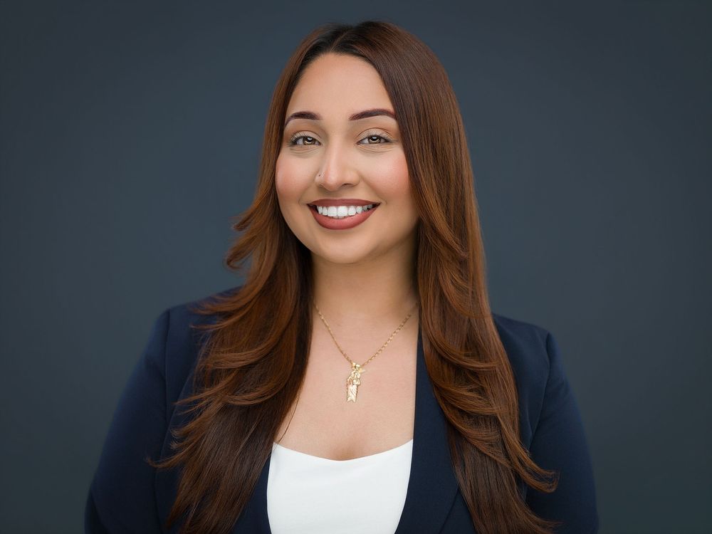 Professional headshot of a smiling young woman with long brown hair wearing a navy blazer and gold necklace.