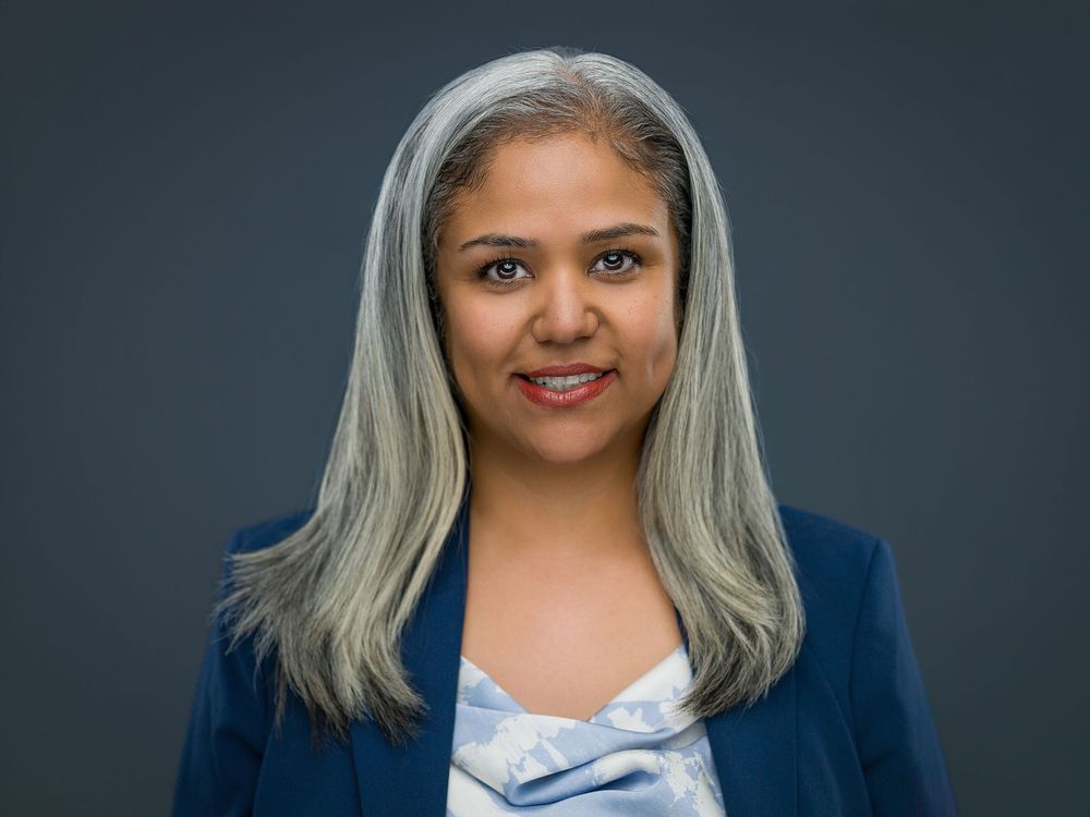 Professional woman with gray-streaked hair wearing a blue blazer, smiling confidently against a dark background.