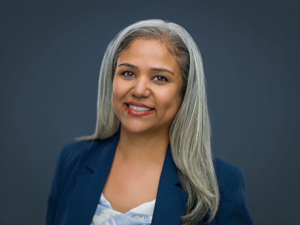 Professional woman with long gray hair wearing a blue blazer, smiling confidently against a dark gray background.