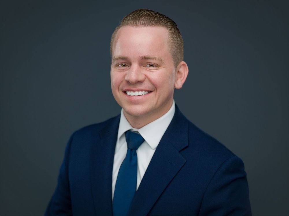 Professional man in navy suit and blue tie smiling confidently against a dark gray background.
