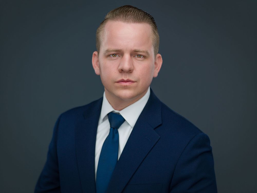 Professional headshot of a young man in a navy blue suit and blue tie against a dark gray background.