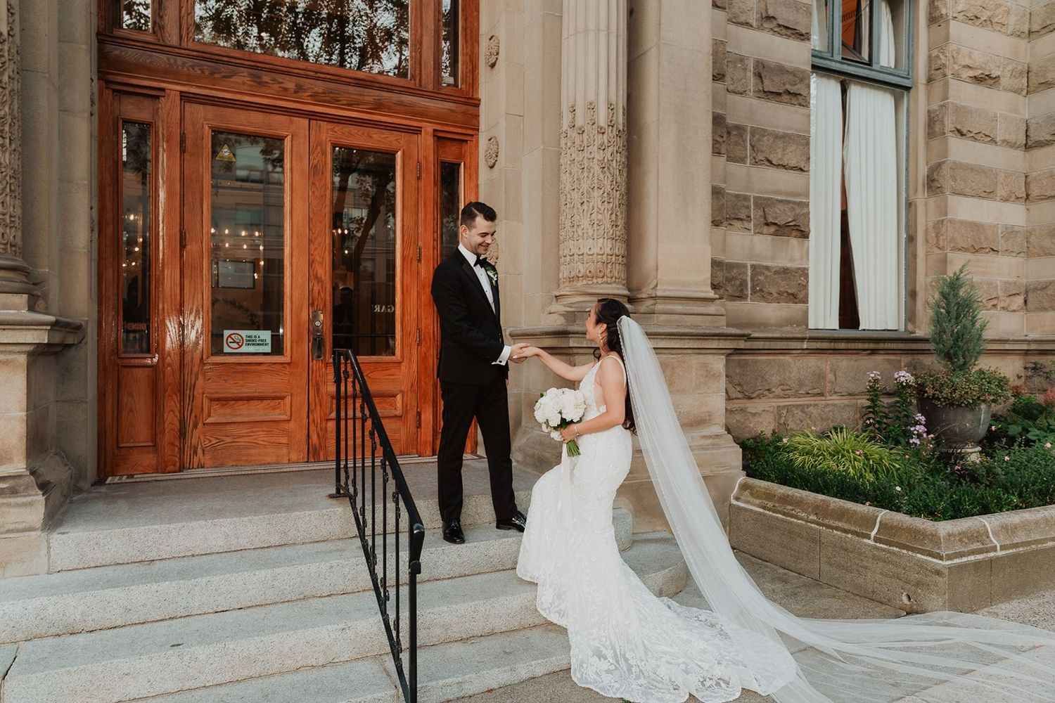 Bride and groom share a romantic first look on stone steps outside an elegant historic building with ornate wooden doors.