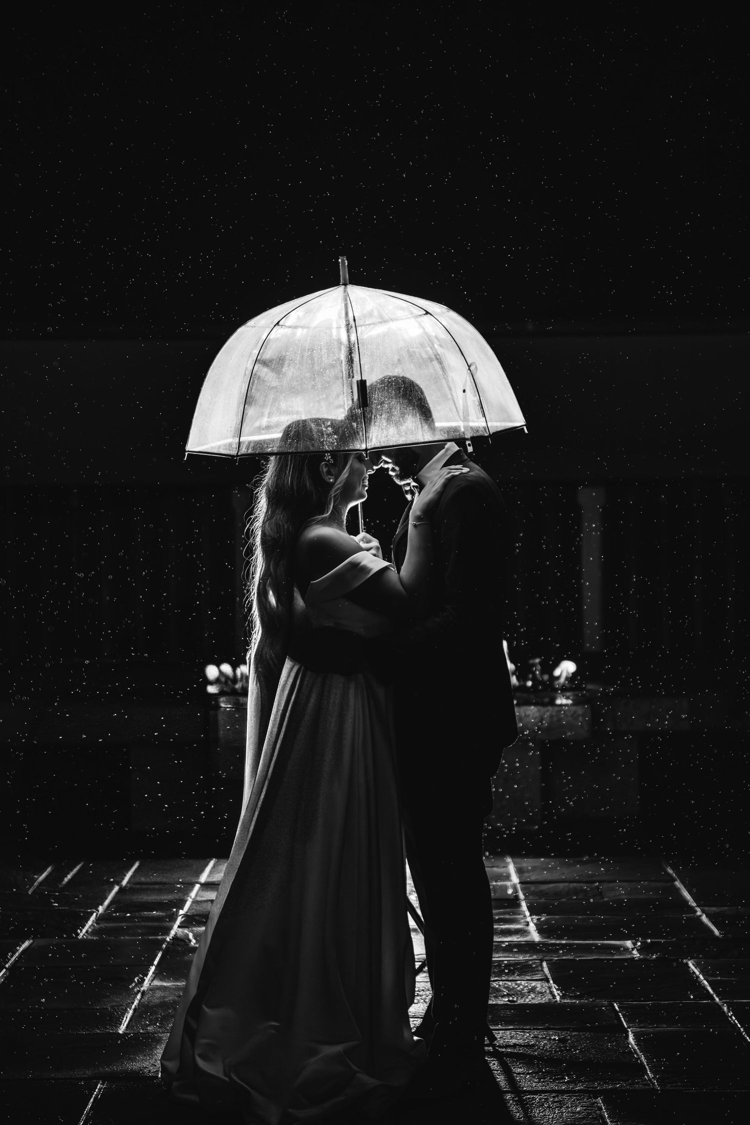 Bride and groom share a kiss under a clear umbrella in a romantic black and white rainy night photo.